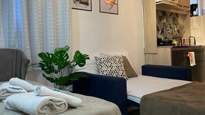A modern hotel room with a bed, a small sofa, a desk, and a kitchenette.