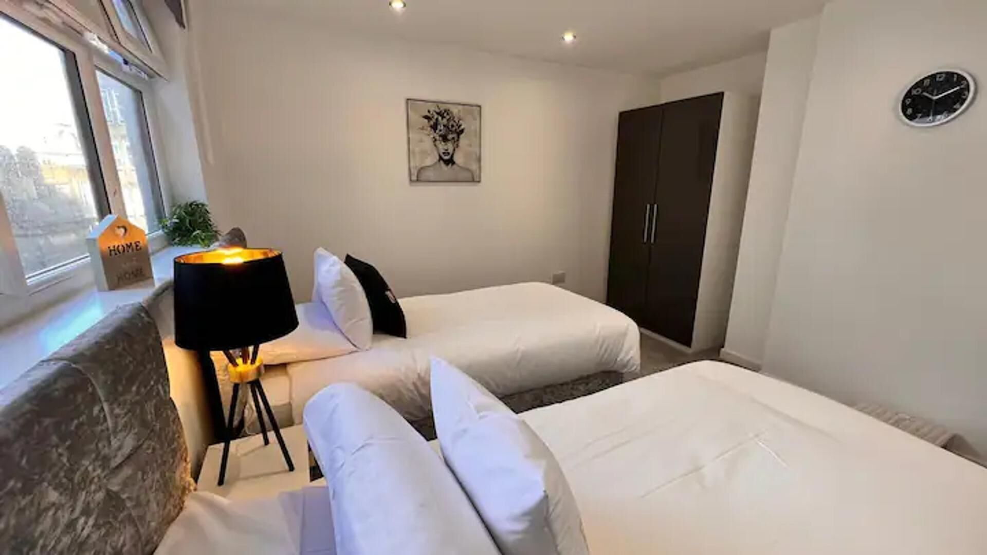 A bedroom with two beds, a bedside table with a lamp, a painting on the wall, and a wardrobe.