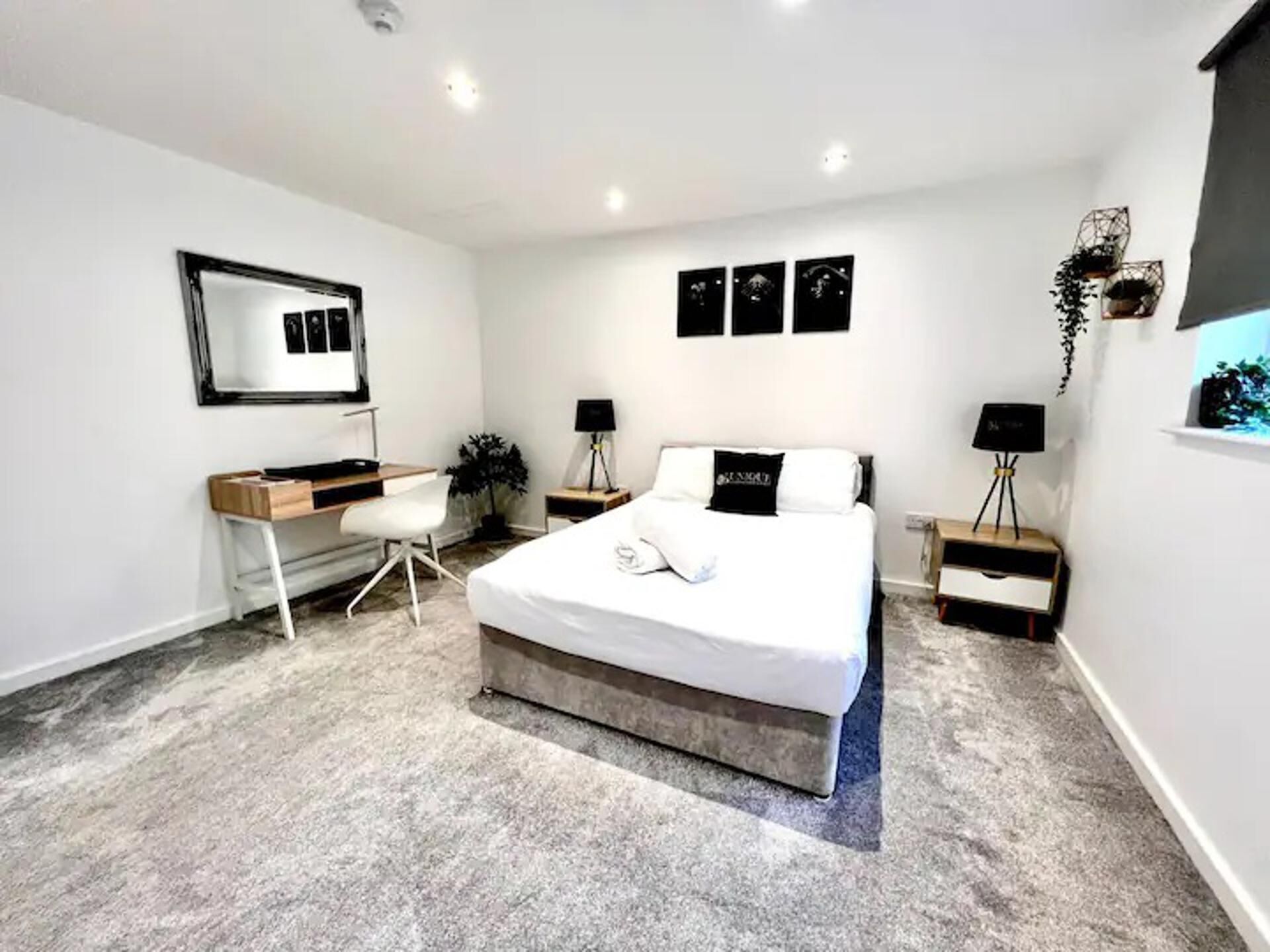 A modern bedroom with a large bed, a desk, and a chair. There are three wall-mounted picture frames and a mirror.