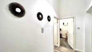 Un couloir aux murs blancs et au plancher en bois, qui mène à une pièce contenant une chaise et une table.