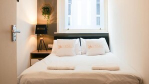 Un lit bien fait, avec du linge de lit blanc et des oreillers, une lampe de chevet et une fenêtre avec des stores.