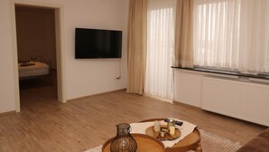 A modern living room with a flat-screen TV mounted on the wall, a wooden coffee table with a candle and remote, and a bed in the adjacent room. - City Apartment With Waterbed (Essen)