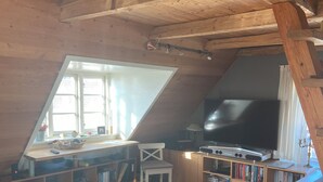 Et hyggeligt loftsværelse med skråvægge, et stort fjernsyn, bogreoler, et skrivebord og en behagelig seng.