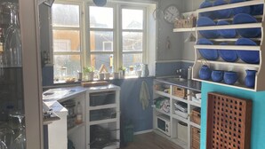 Et lille køkken med trægulv, et vindue med gardiner, og en blå væg med hylder med blå skåle og en træreol.
