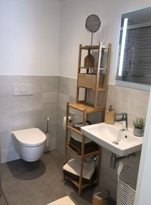 A compact bathroom with a toilet, a wooden shelf unit, a sink with a mirror, and a small trash bin. - Pleasant Apartment With Terrace (Ohningen)