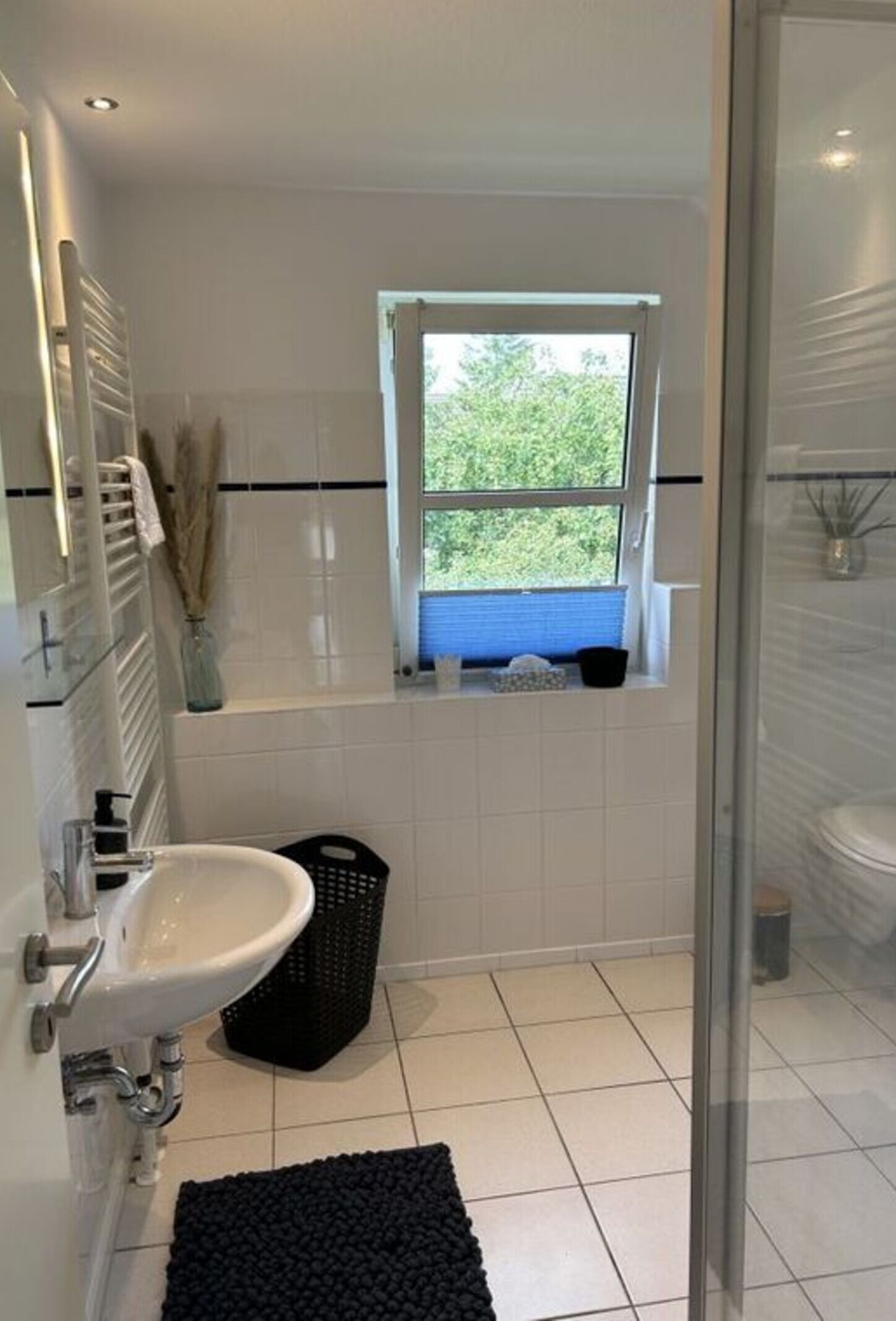 A modern bathroom with a white sink, a glass shower enclosure, a window with a view of greenery, and a black bath mat.