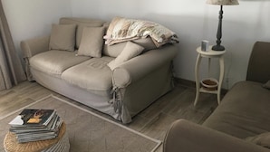 Un sofá color beige con una manta de flores, una mesita con una lámpara y una cesta tejida en el suelo.