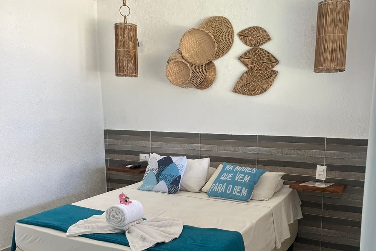 A neatly made bed with a teal blanket, a pillow with a blue and white pattern, and a pillow with a blue text. There are woven baskets on the wall and two hanging lamps.