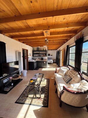 A spacious living area with a wooden ceiling, a sofa, a TV, and a kitchen in the background.