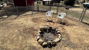 A fenced outdoor area with a fire pit, two white chairs, a red barn, and a mountainous backdrop. - Alarcon Ranch  (Weldon)