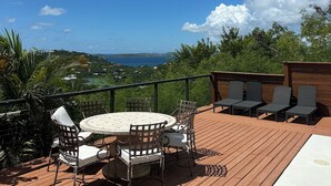 Eine Holzterrasse mit rundem Tisch und Stühlen, mit Blick auf eine reizvolle Landschaft mit Bäumen und Meer.