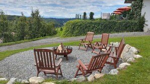 A circular outdoor seating area with wooden chairs and a fire pit, surrounded by a gravel path and greenery, with a view of a building and hills in the background. - Apartment in Schwalbenhof Farm (Sankt Stefan im Rosental)