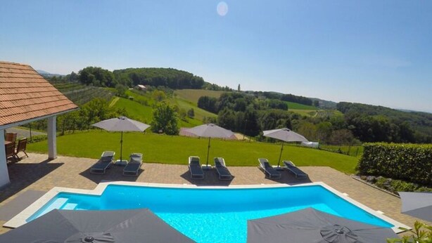 A swimming pool with lounge chairs and umbrellas, surrounded by a landscaped garden and a view of rolling hills. - Apartment in Schwalbenhof Farm (Sankt Stefan im Rosental)