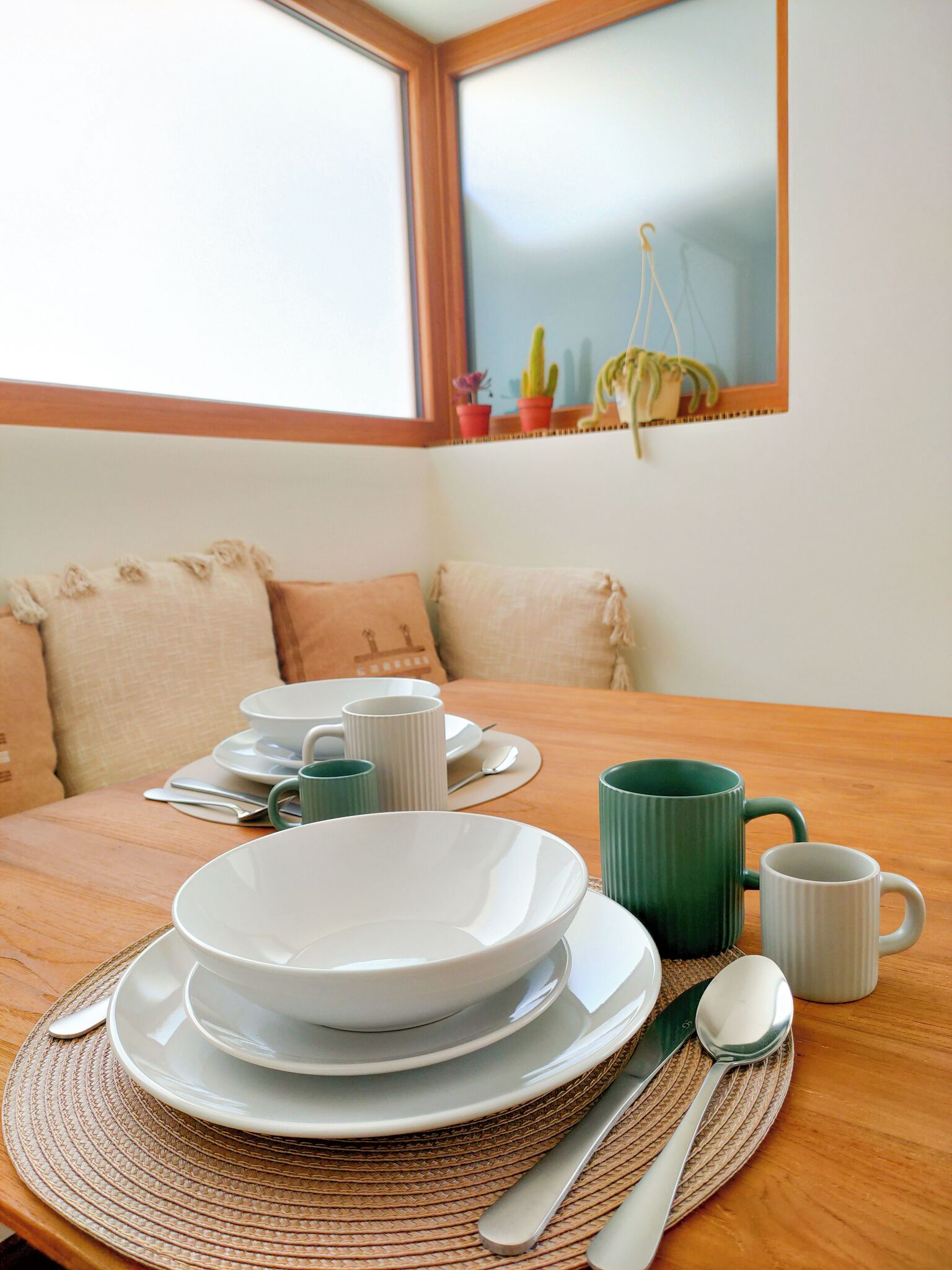 Une table à manger dressée avec des assiettes, des bols et des tasses, et une fenêtre donnant sur un cactus.