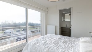 Een moderne slaapkamer met een groot raam, een bed, een dressoir en een spiegel.