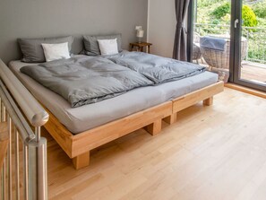 Un dormitorio con estructura de cama de madera, ropa de cama gris, una mesita de noche pequeña con una lámpara y vistas al exterior a través de un ventanal corredero.