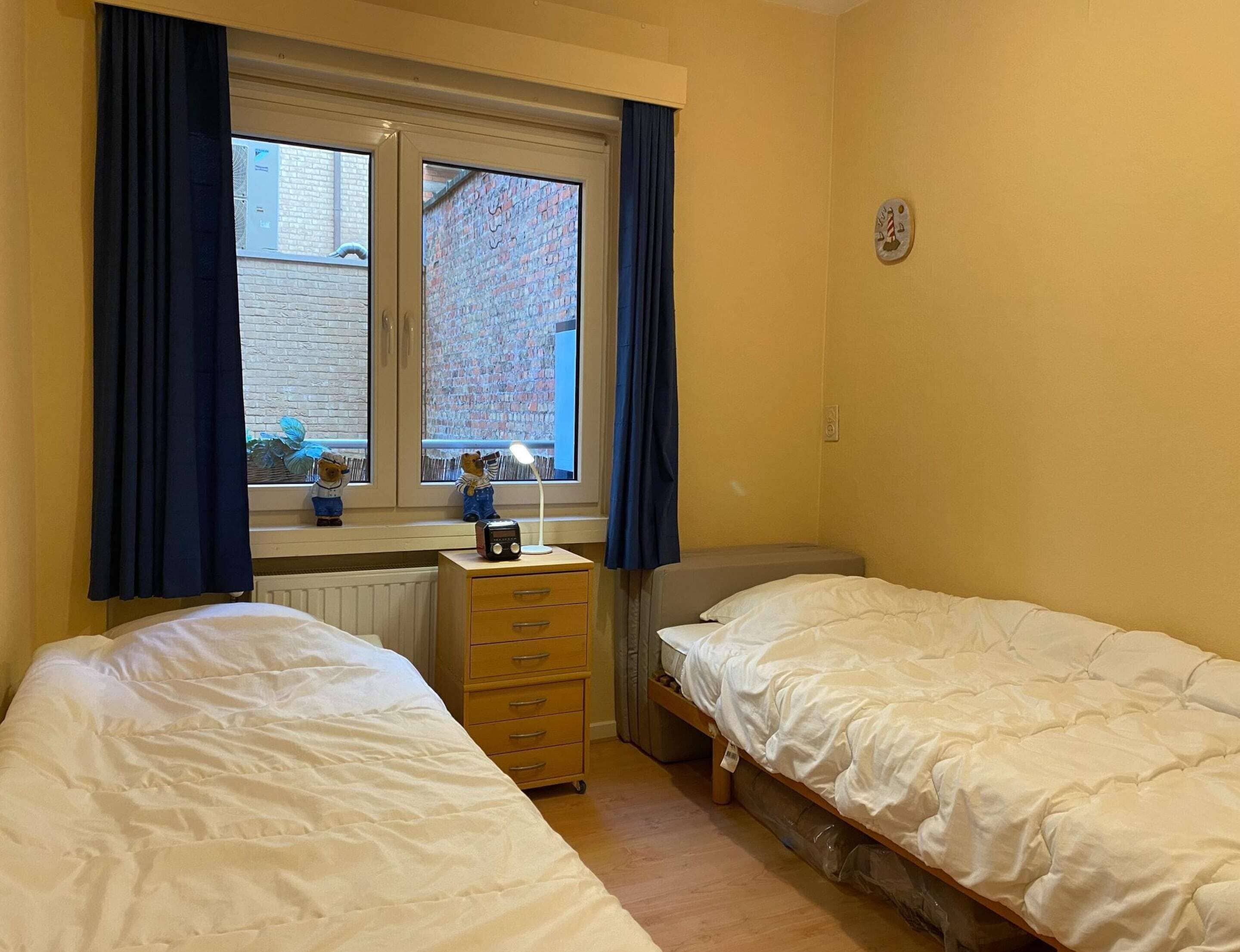A bedroom with two single beds, a desk with a lamp, a radiator, and a window with curtains.