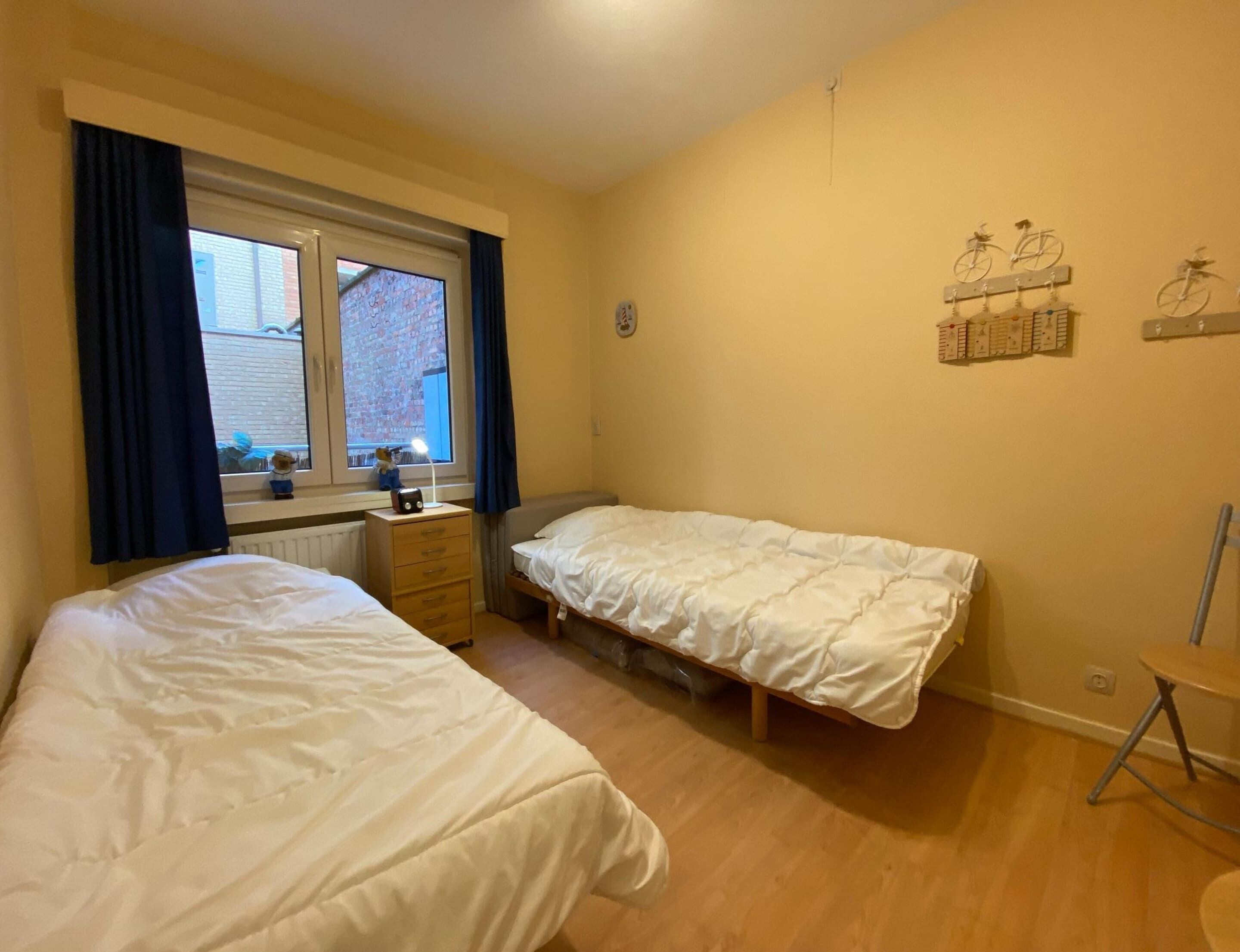 A bedroom with two single beds, a dresser, a desk with a lamp, and a window with curtains.