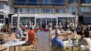 An outdoor dining area with tables and chairs, a building with balconies in the background, and a sign that reads "RESTAURANT". - Apartment in Nieuwpoort With Views (Nieuwpoort)