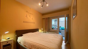 A bedroom with a large bed, bedside lamps, a small table, and a window with a view of the outside balcony. - Apartment in Nieuwpoort With Views (Nieuwpoort)