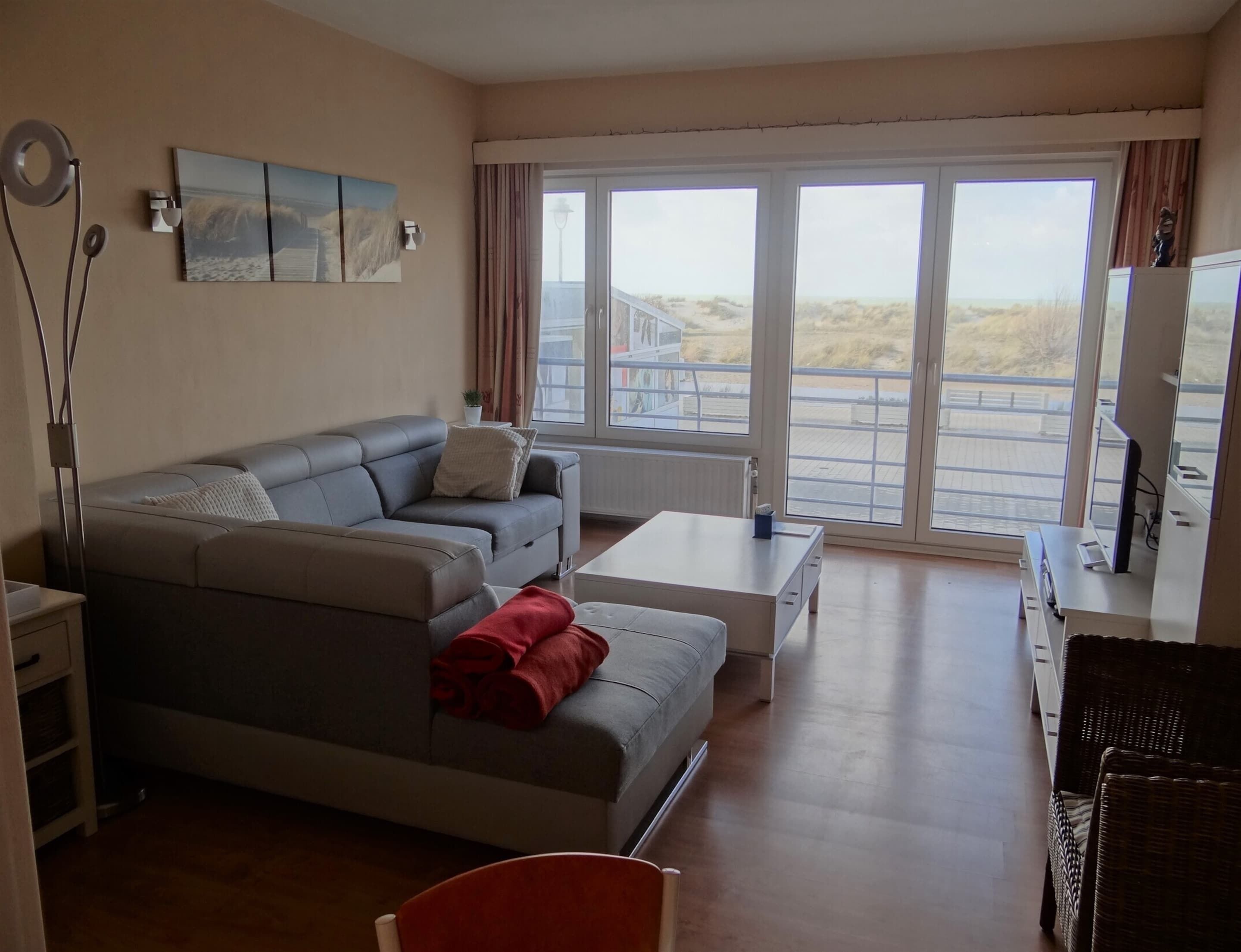 A modern living room with a grey sectional sofa, a small white coffee table, and a red chair. There is a large window with a view of the beach and a balcony.