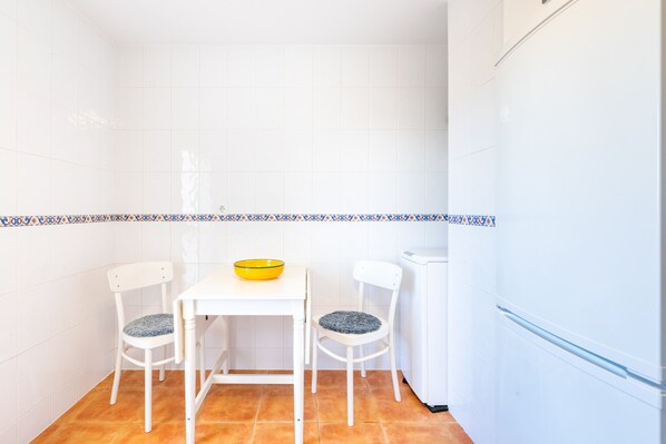 Une petite cuisine aux murs recouverts de carreaux blancs, avec un réfrigérateur blanc, une table blanche et deux chaises blanches dotées de coussins bleus.