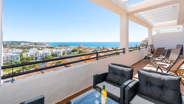 Un balcon avec vue sur la mer, agrémenté de fauteuils et d’une table en osier, ainsi qu’une bouteille de vin.