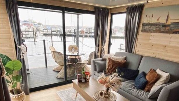 Una sala de estar moderna con un sofá gris, una mesa de centro de madera y vistas a un puerto deportivo a través de amplios ventanales.