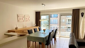 A dining area with a long table, chairs, a wooden bench, and a view of the outside through large windows. - Apartment in Nieuwpoort With Ocean Views (Nieuwpoort)