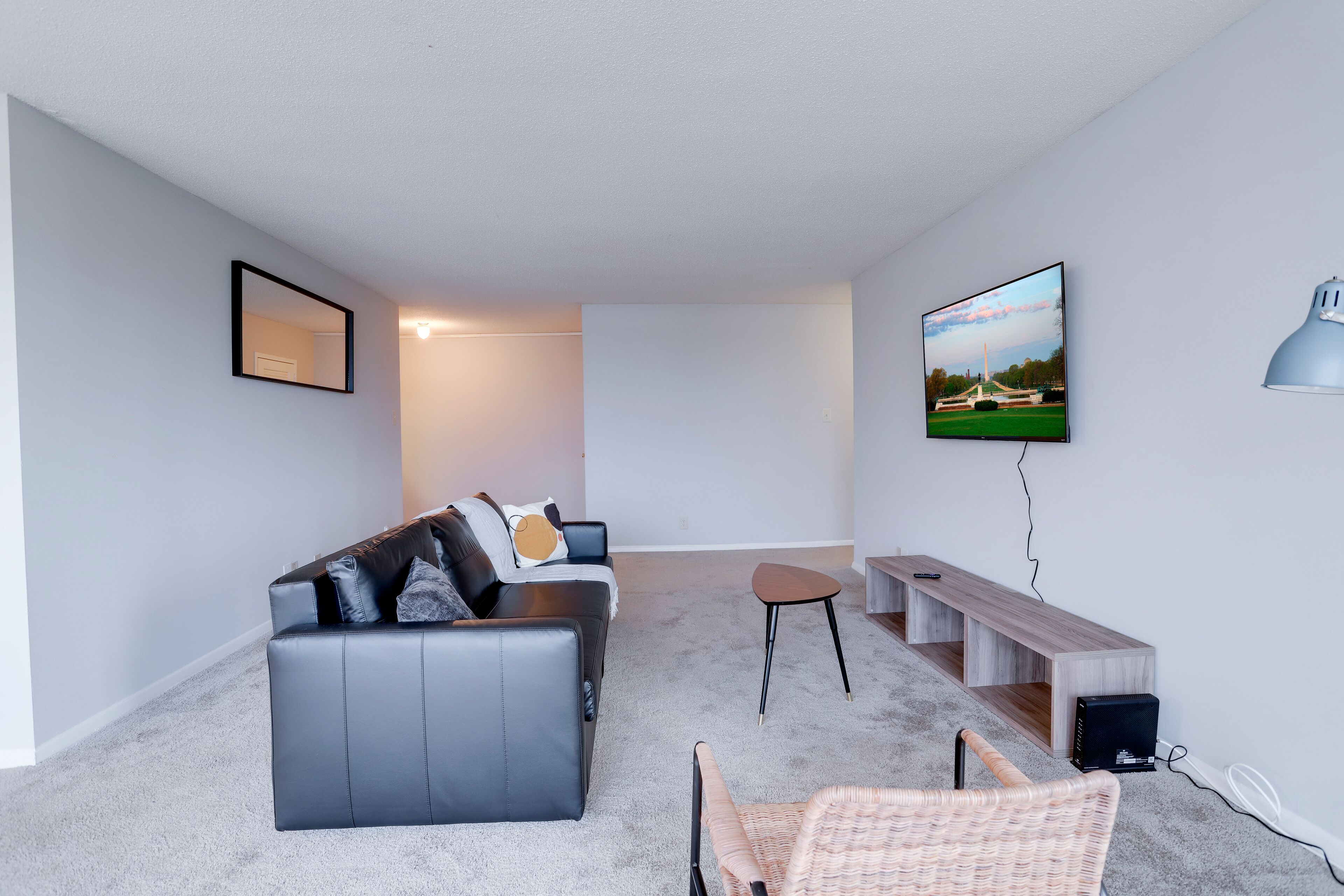 A modern living room with a black leather sofa, a small round table, a wicker chair, a TV mounted on the wall, and a wooden bench.