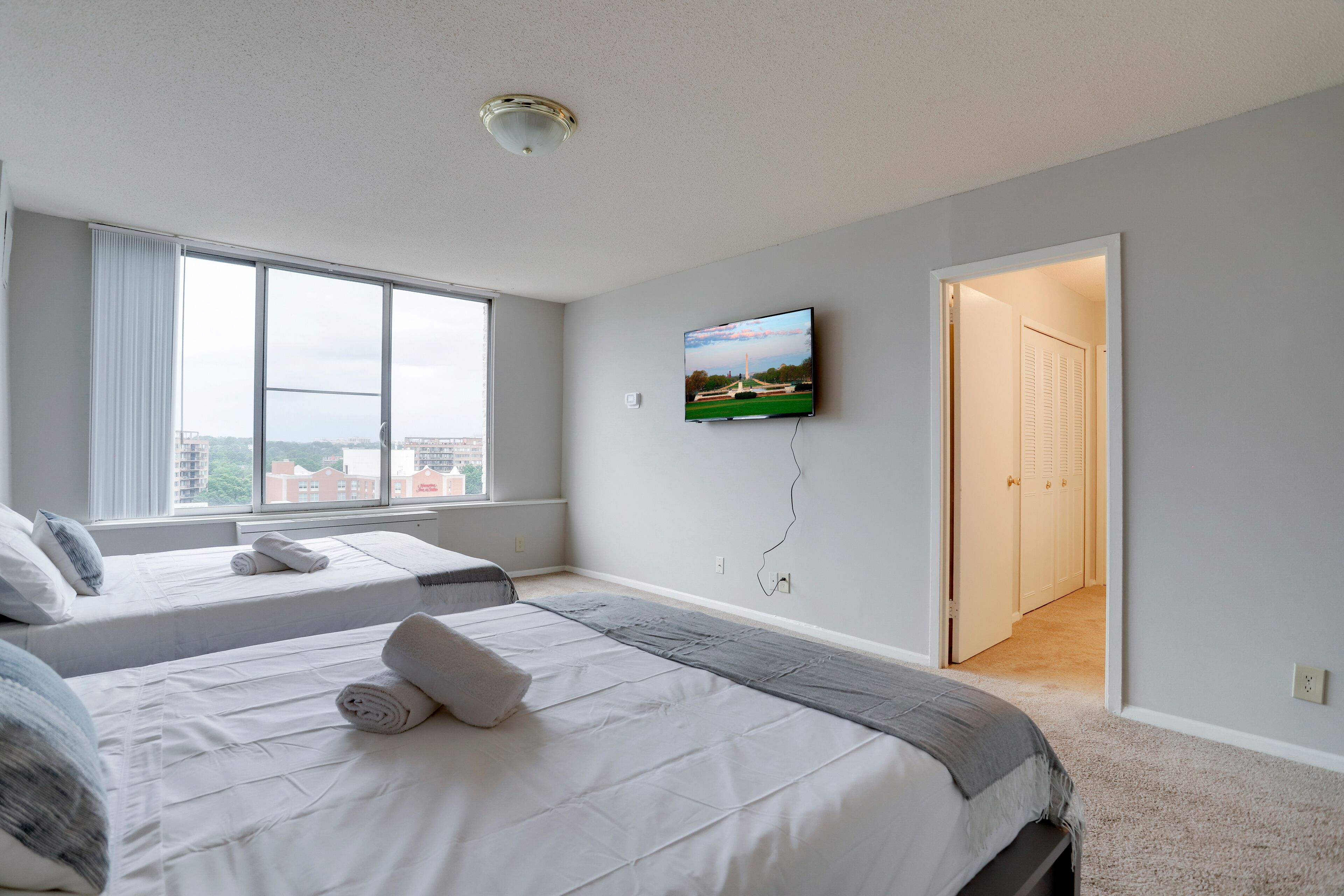 Quarto de hotel com duas camas, televisão na parede e vista para a cidade através de grandes janelas.