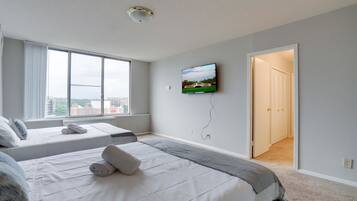 Quarto de hotel com duas camas, televisão na parede e vista para a cidade através de grandes janelas.