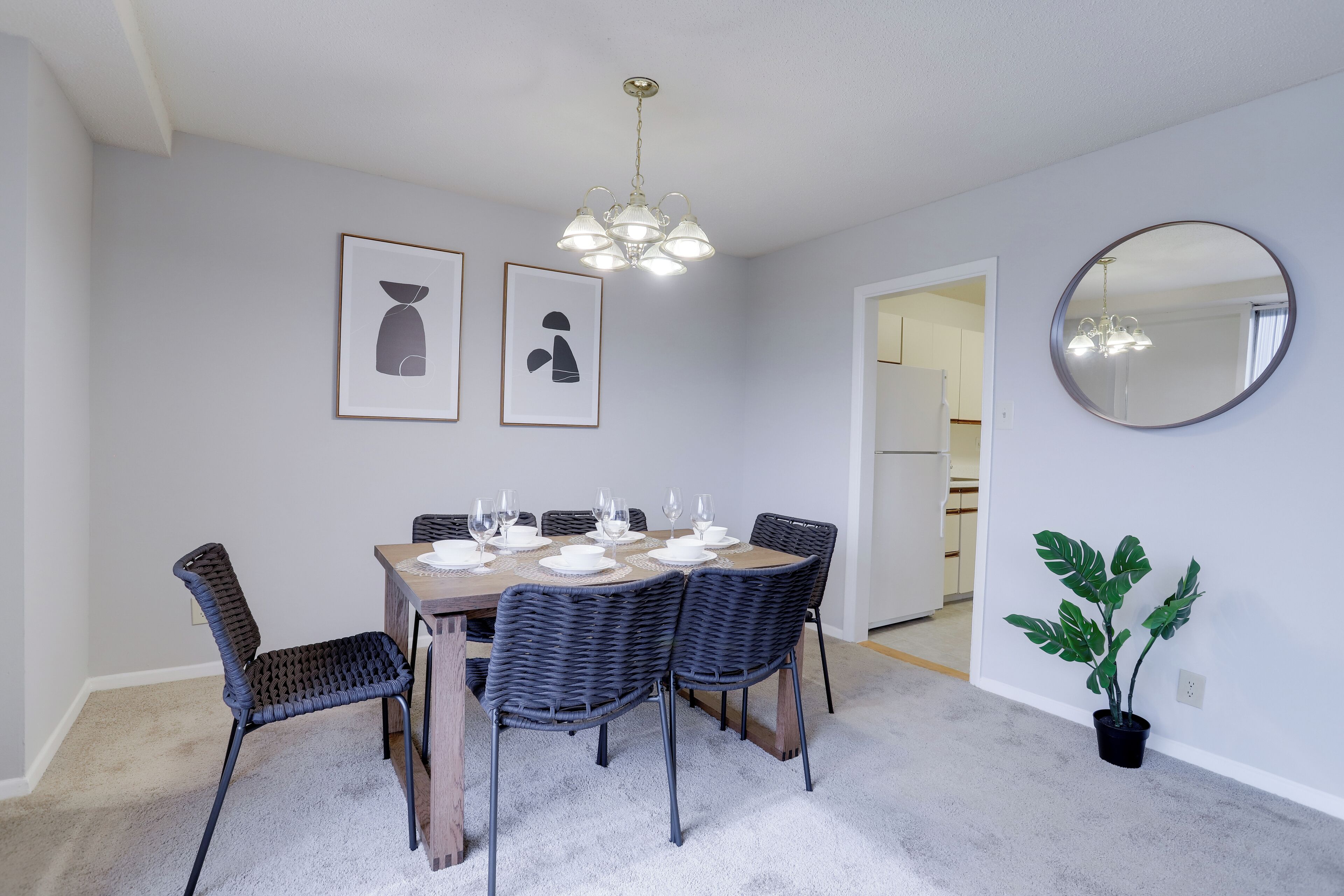 Un comedor con una mesa de madera dispuesta para seis personas, rodeada de sillas de mimbre oscuro. Se observa una araña sobre la mesa y una planta en maceta a la derecha.