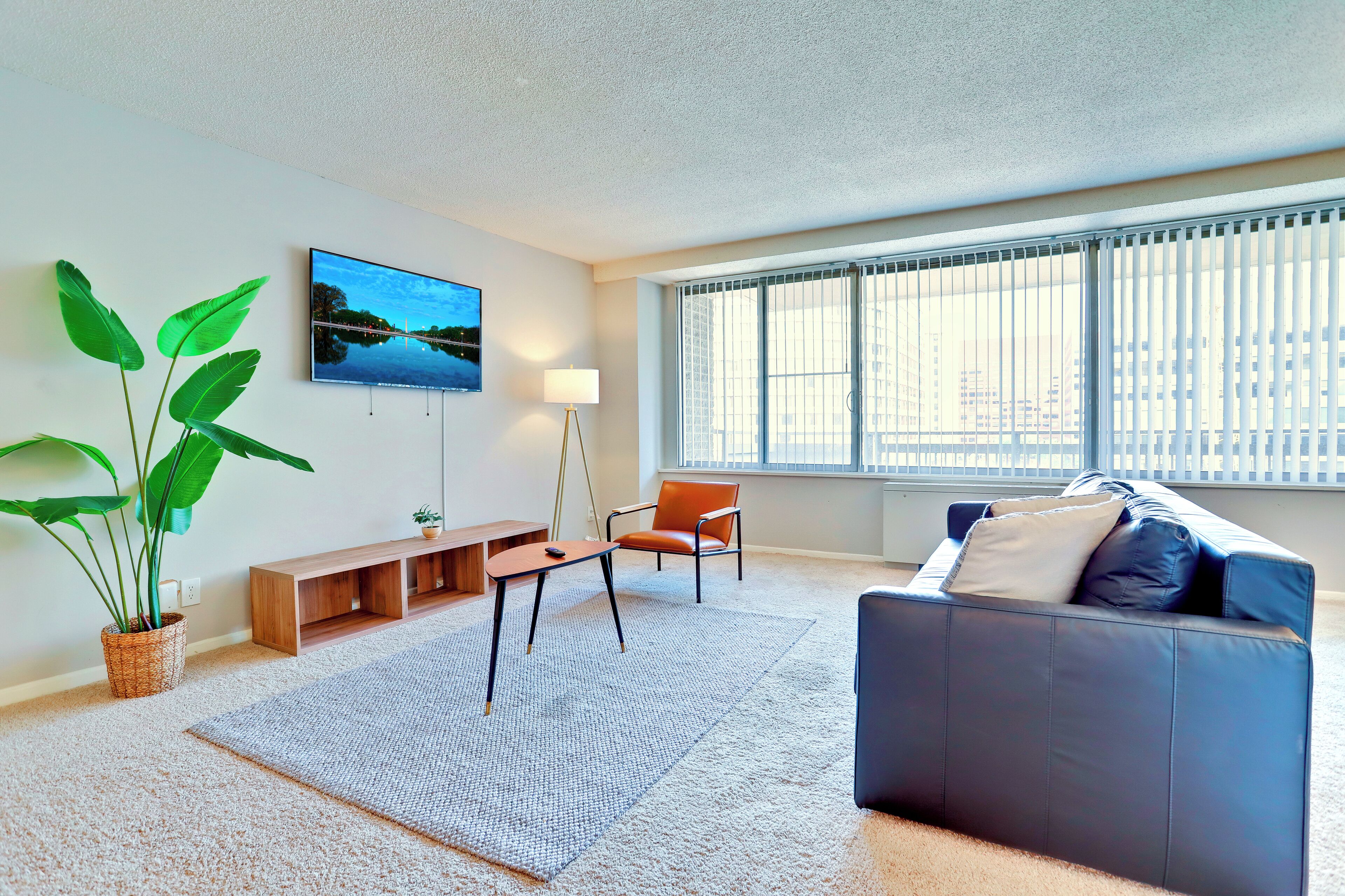 Una sala de estar moderna con un sofá gris, una mesa de centro, un televisor, una planta y un ventanal con persianas.