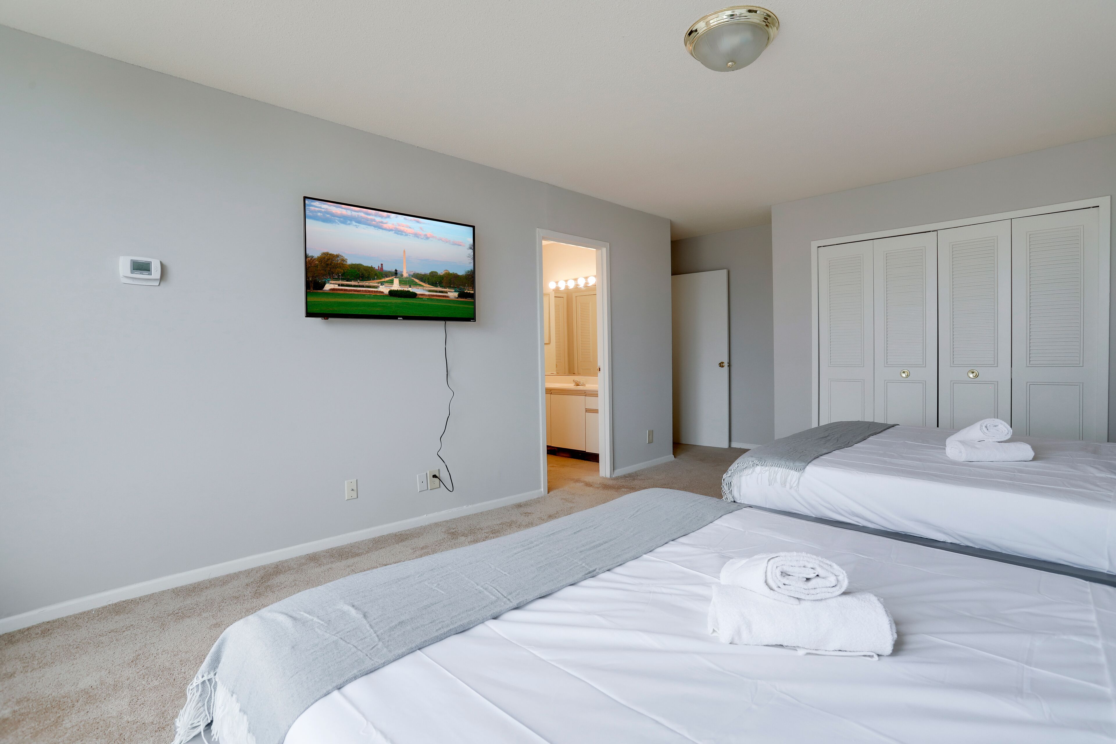A hotel room with two beds, a TV on the wall, a hallway leading to a bathroom, and a closet with louvered doors.