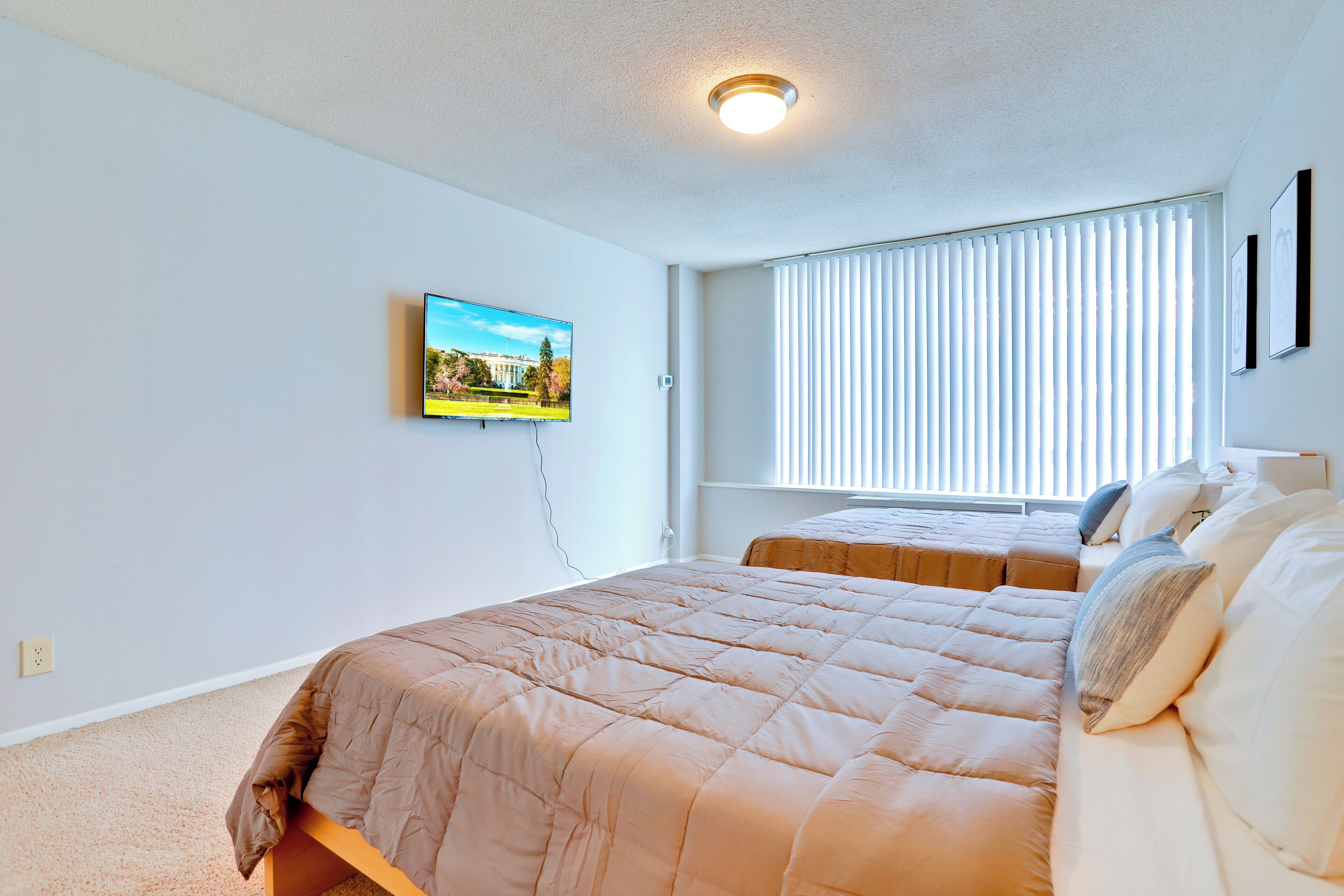 A bedroom with a large bed, a smaller bed, a TV on the wall, and a window with blinds.