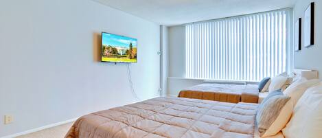 A bedroom with a large bed, a smaller bed, a TV on the wall, and a window with blinds.