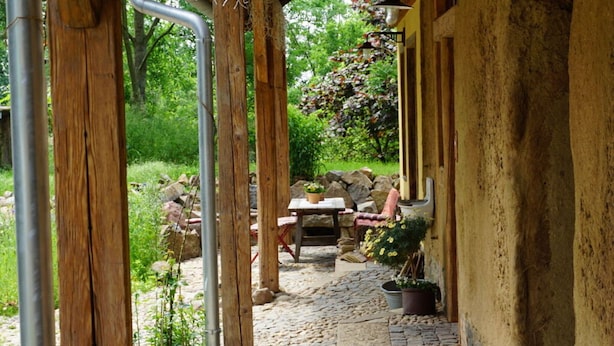 Une terrasse extérieure rustique, dotée d’un sol en pierre, de piliers en bois et d’une table avec des chaises.