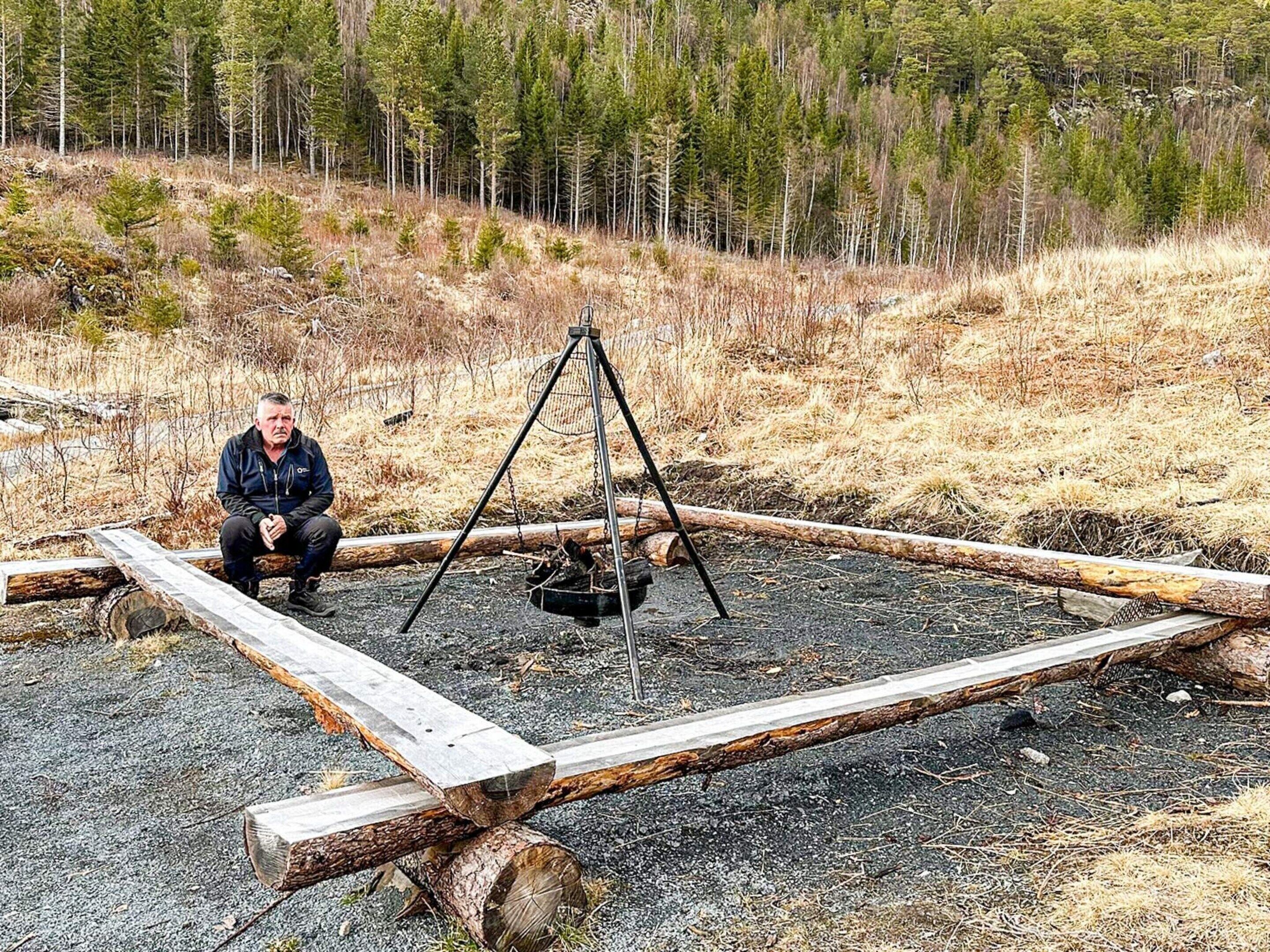 Uma pessoa sentada ao lado de uma estrutura metálica com assento de balanço, cercada por toras e uma área arborizada.