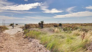 A coastal landscape with a sandy path, sparse vegetation, and a clear sky.