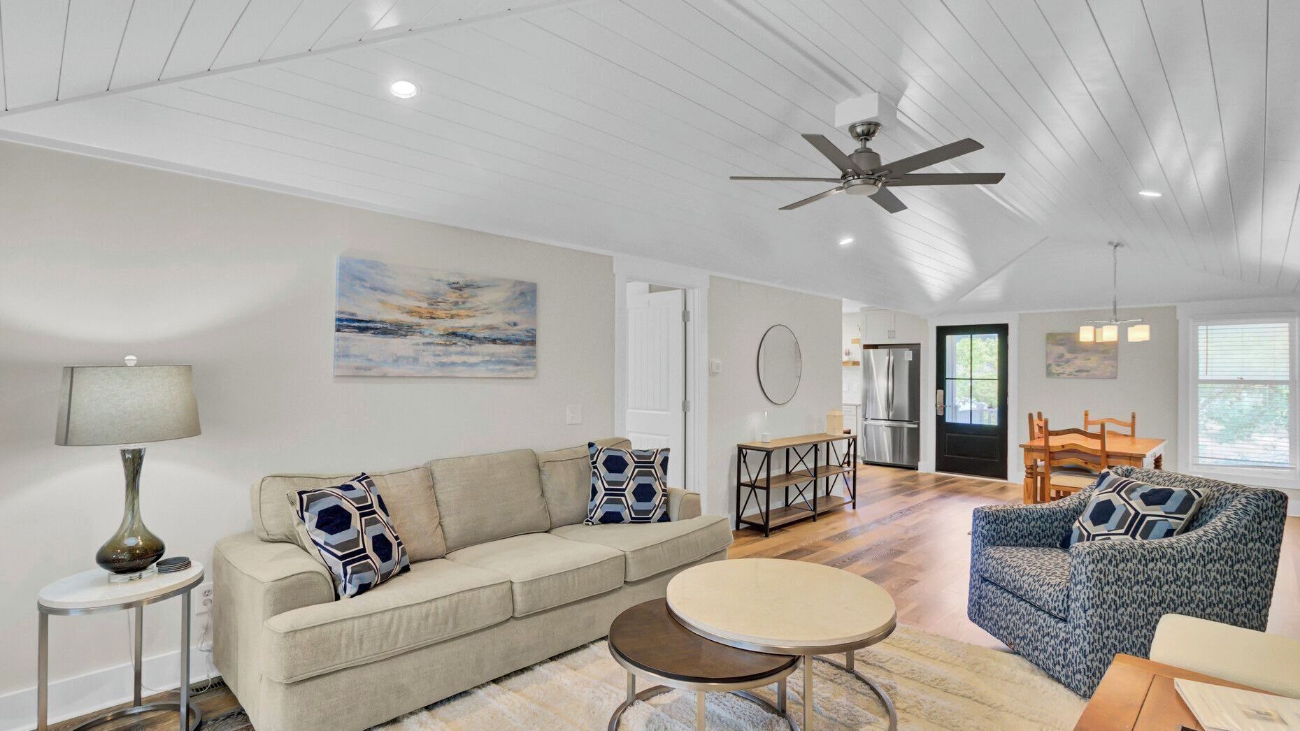 A spacious living area with a sofa, coffee table, and patterned armchair, a ceiling fan, and a dining area with a table and chairs.