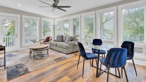 A spacious room with a ceiling fan, large windows with blinds, a grey sofa, blue chairs, and a round coffee table.