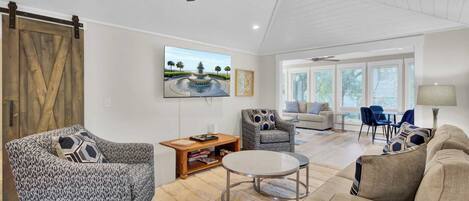 A spacious living room with a ceiling fan, a large TV mounted on the wall, and a view of the outdoors through large windows.