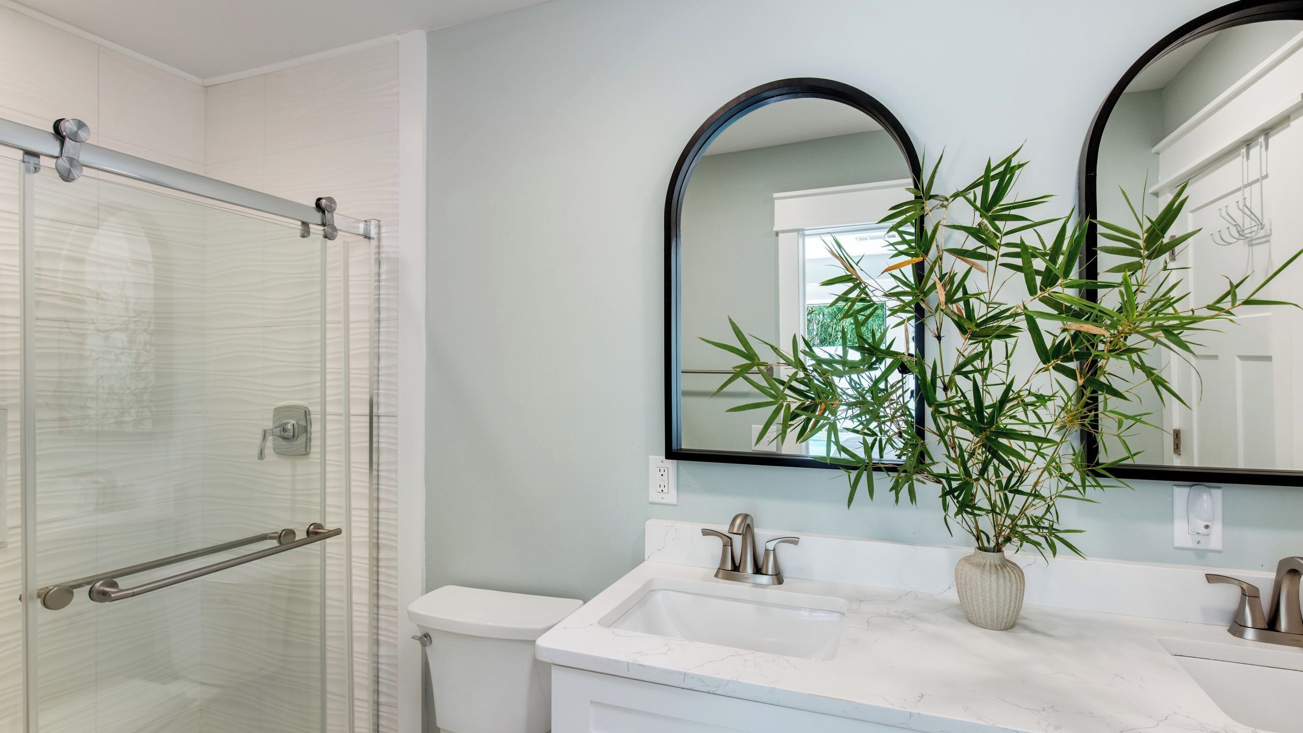 A bathroom with a glass shower enclosure, a white toilet, a sink with a marble countertop, and a mirror with a bamboo plant.