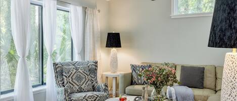 Un salon comprenant un fauteuil à motifs, une table basse ronde avec des livres et un vase, un canapé avec des coussins et une lampe sur une table d’appoint.