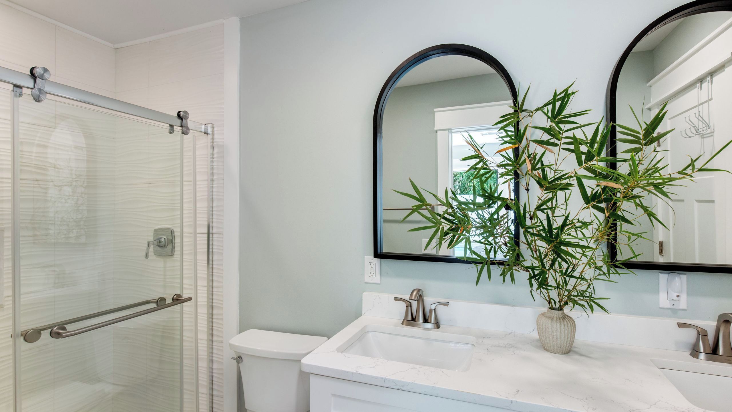 A bathroom with a glass shower enclosure, a toilet, a sink with a mirror, and a potted plant.