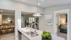 A modern kitchen with a built-in sink, a dining area with a round mirror, and a bedroom with a bed and nightstand. - Hilton Hideaway by Avantstay Private Pool, Walk to Beach (Hilton Head Island)