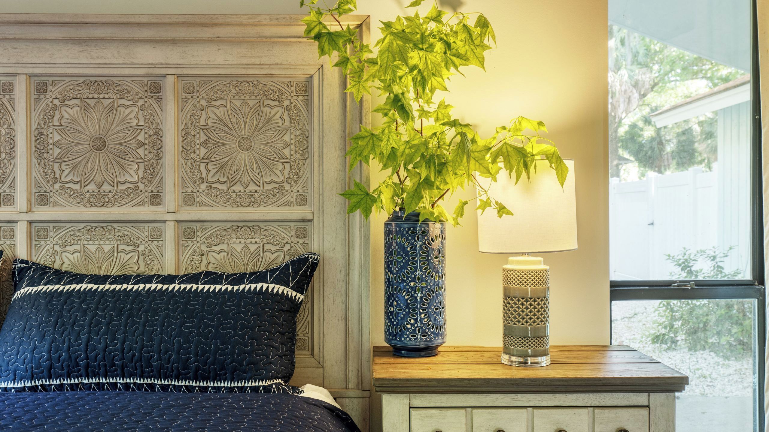 Un dormitorio con un cabecero decorativo, una cama con una colcha estampada oscura, un jarrón azul con una planta y una lámpara de noche sobre una mesita de madera.