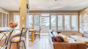 A spacious living area with a wooden floor, a brown leather sofa, a dining table with chairs, and large windows offering a view of trees and a garage. - Sunvail by Avantstay 10min Walk to Ski Lifts! (Vail)