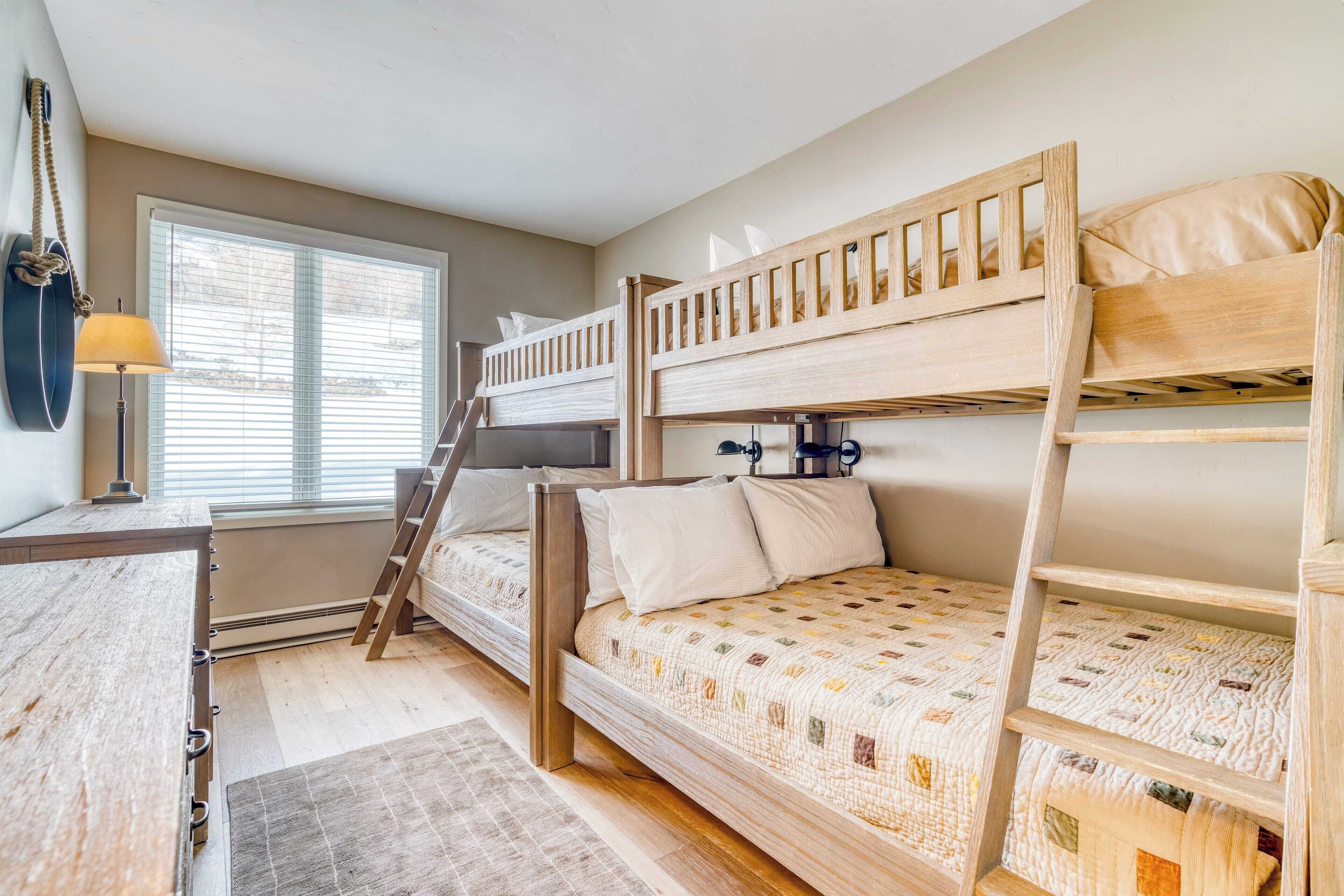 A bunk bed room with a ladder, a desk with a lamp, and a window with blinds.